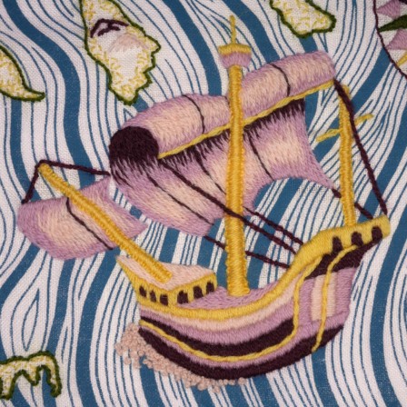 Gros plan sur une broderie de carte marine : un bateau style caravelle avec 2 voiles et 3 mats. Il brodé en différentes nuances de rose et en jaune. La couleur la plus foncée est assortie aux 2 roses clairs mais elle est très foncée, presque noire.