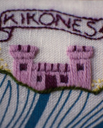 Gros plan sur une broderie de carte marine. Sur la côte brodée en vert et jaune, des remparts brodés en rose et pourpre. Au dessus un phylactère en forme de parchemin avec écrit &amp;quot;Kikonès".