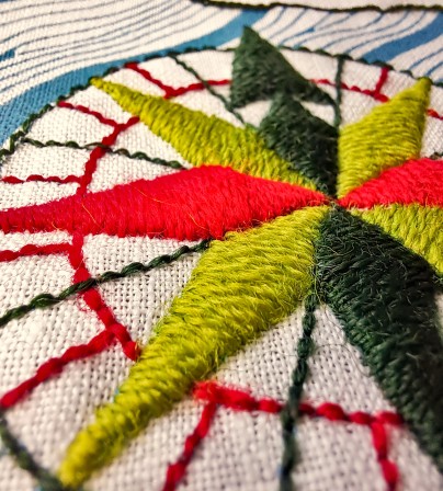 Gros plan sur la moitié de la rose des vents. Elle est brodée avec des flèches vert clair, rouge et vert foncé dans un point de remplissage au rendu très lisse.