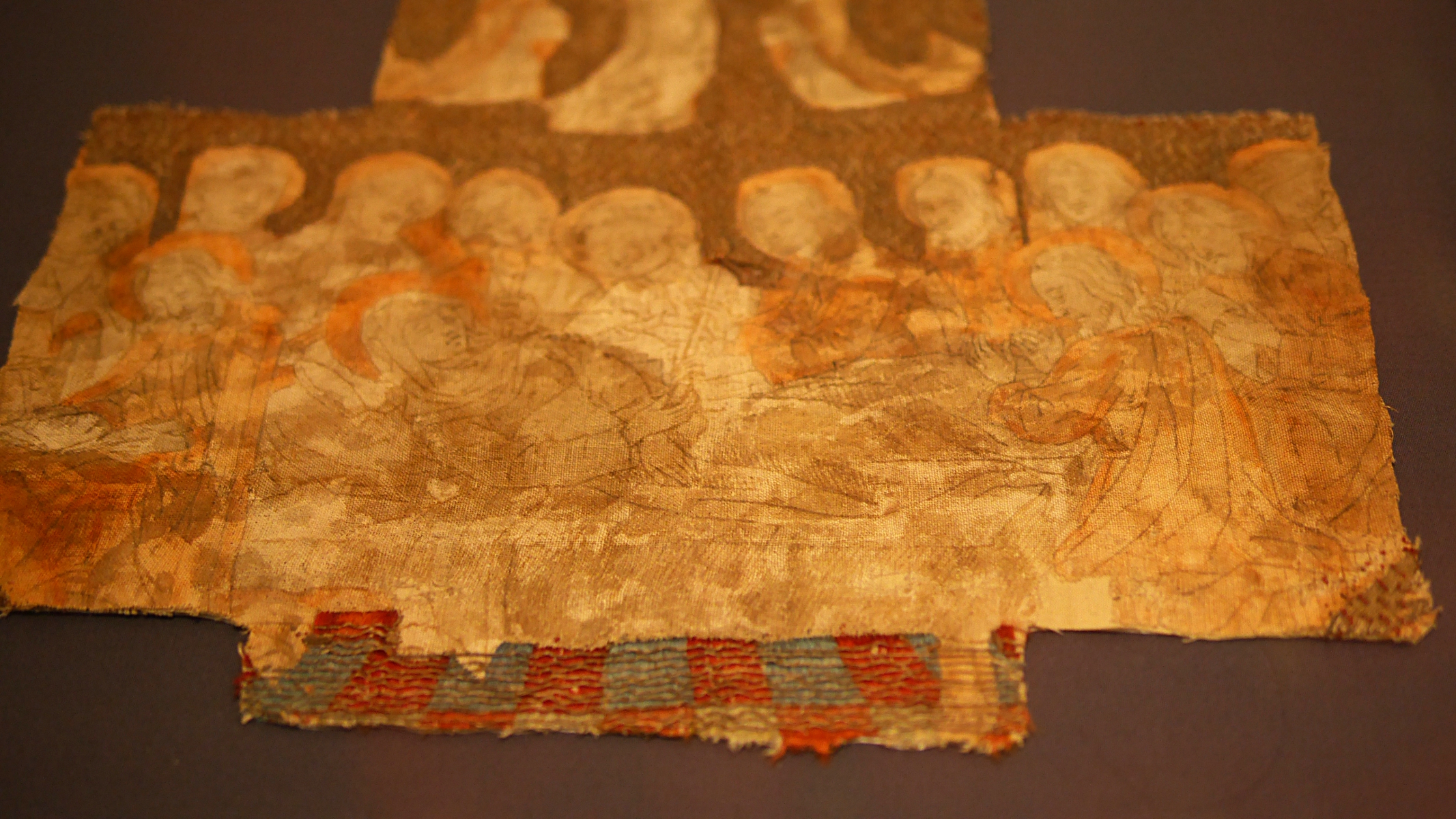 Détails d'une broderie non terminée de la fin du moyen-âge. Il s'agit de la vierge allongée sur son lit avec une foule qui l'entoure en train de prier. Seul le fond est brodé et on voit le motif tracé à la main à l'encre et des zones peintes en rouge.