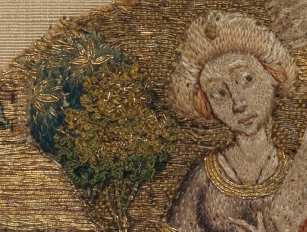 Détail d'une broderie médiévale au fil d'or et de soie colorée. On distingue à côté de la tête d'une femme un arbre brodé dont le feuillage est réalisé avec des points entortillés.