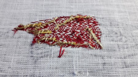 Détail d'une broderie vue sur l'envers. On distingue une zone brodée, elle est recouverte de fils rouges qui forment un motif géométrique un peu flou. Par dessus, on voit le bout des fils d'or qui sont fixés sur la zone brodée avec des points.