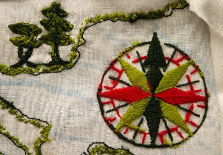 Envers d'une broderie en laine colorée, on distingue sur la droite une rose des vents et sur la gauche des arbres. On voit aussi une ligne de côte avec des petits points verts tout le long.