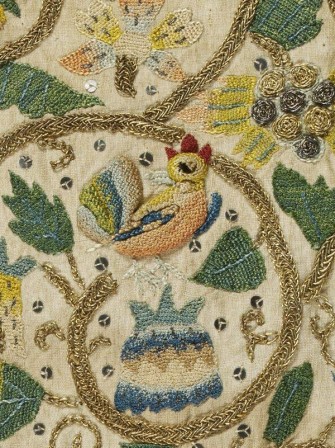 Détail d'une broderie sur toile écrue avec un décors végétal et un coq coloré au centre. La broderie est réalisée avec des fils colorés et les tiges sont brodées au fil d'or avec un point tressé