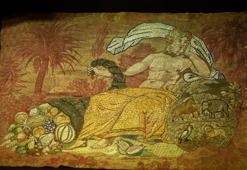 Broderie sur une toile de Jouy imprimée en rouge représentant un dieu antique (Zeus) assis à côté d'une grande corne d'abondance qui déborde. Le personnage et la corne sont brodés, pas l'arrière plan.