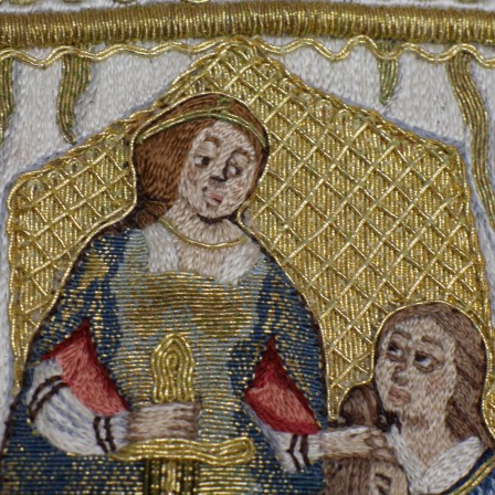 Détail de la broderie du personnage de Judith. Elle est brodée au fil de soie coloré. Sa robe est brodée au fil d'or recouvert de différentes nuances de fils bleus. Elle porte des bijoux brodés au fil d'or. Son regard est dirigé vers le bas, vers sa servante.